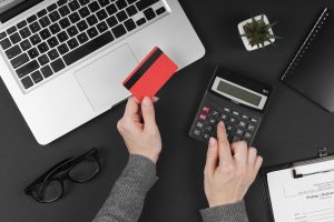 A top-down view of a workspace featuring a silver laptop, a black calculator, a red credit card, and a pair of black eyeglasses on a dark background. A person wearing a gray sweater holds the credit card in one hand while using the calculator with the other. Nearby, there is a small potted succulent plant, a black spiral notebook, and a clipboard with a document labeled "Pickup / Delivery." The setup suggests an online shopping, financial planning, or budgeting scenario.