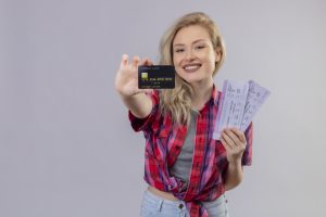 A young woman with blonde hair and a bright smile is posing against a plain light gray background. She is wearing a red plaid shirt tied at the waist over a gray crop top and light blue jeans. She holds a black credit card in her right hand, extending it towards the camera, while her left hand holds a set of airline tickets. The credit card has visible numbers and the text "CREDIT CARD" on it. She appears excited and ready for travel.