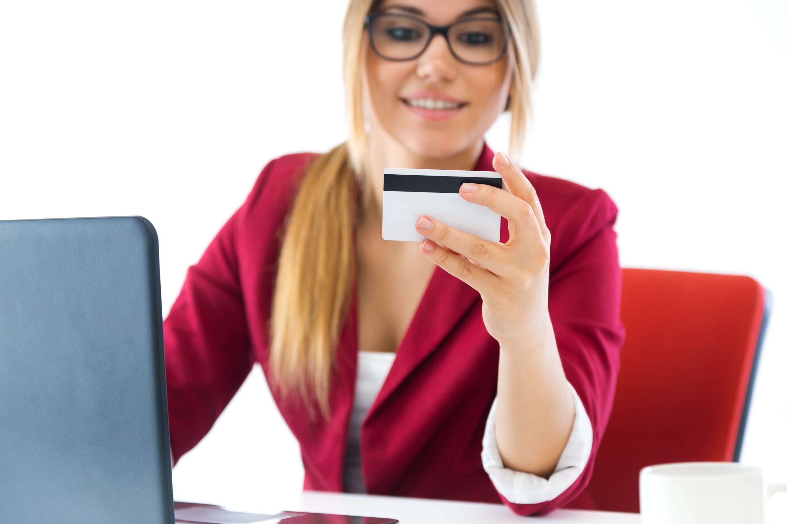 A young woman with long blonde hair, glasses, and light skin is sitting at a desk, holding a credit card while looking at it with a smile. She is wearing a red blazer over a white top. In front of her is an open laptop, and a white coffee cup is placed on the table. The background is bright and minimalistic, suggesting a professional or home office environment. She appears to be making an online purchase or managing her finances.