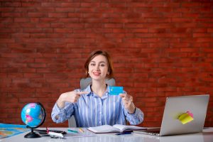 Smiling woman sitting at a desk in front of a brick wall, pointing at a blue credit card she is holding, with travel items like a globe, map, and laptop on the table.