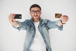A young man in a denim jacket and glasses holding two credit cards, one black and one gold, while smiling at the camera, set against a plain white background.