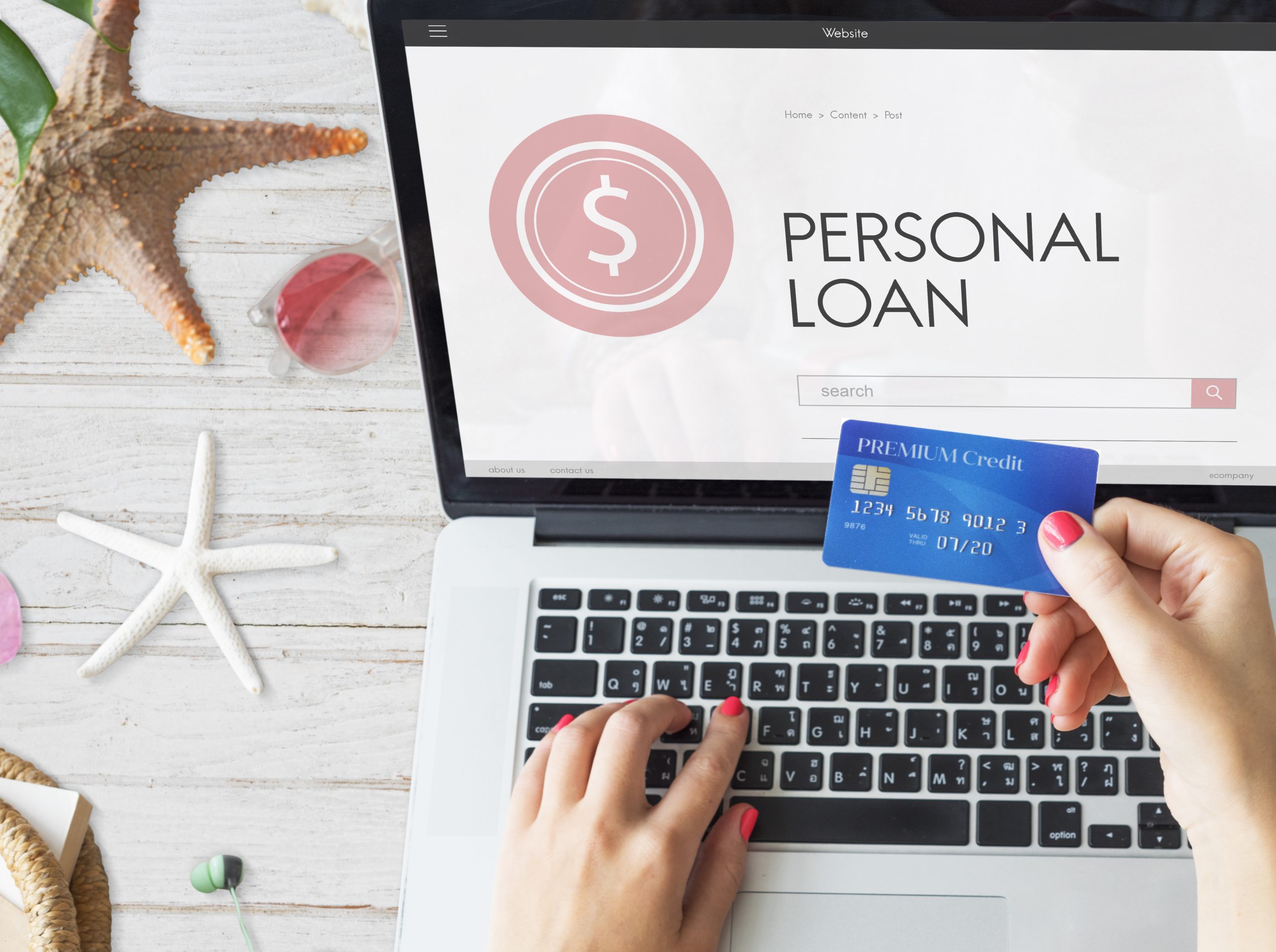 A woman browses a website about personal loans on her laptop while holding a blue premium credit card in one hand. Her pink-painted nails stand out against the silver keyboard, and summer-themed items like starfish, sunglasses, and seashells are scattered on the wooden desk. The image symbolizes financial decision-making in a relaxed setting, highlighting the comparison between credit cards versus personal loans.