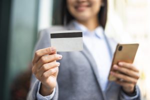 A smiling professional woman in a blazer holds a smartphone in one hand and displays a blank credit card in the other. The business setting and confident demeanor highlight the convenience and rewards offered by credit cards with business travel benefits, such as airline miles, hotel perks, and travel insurance.