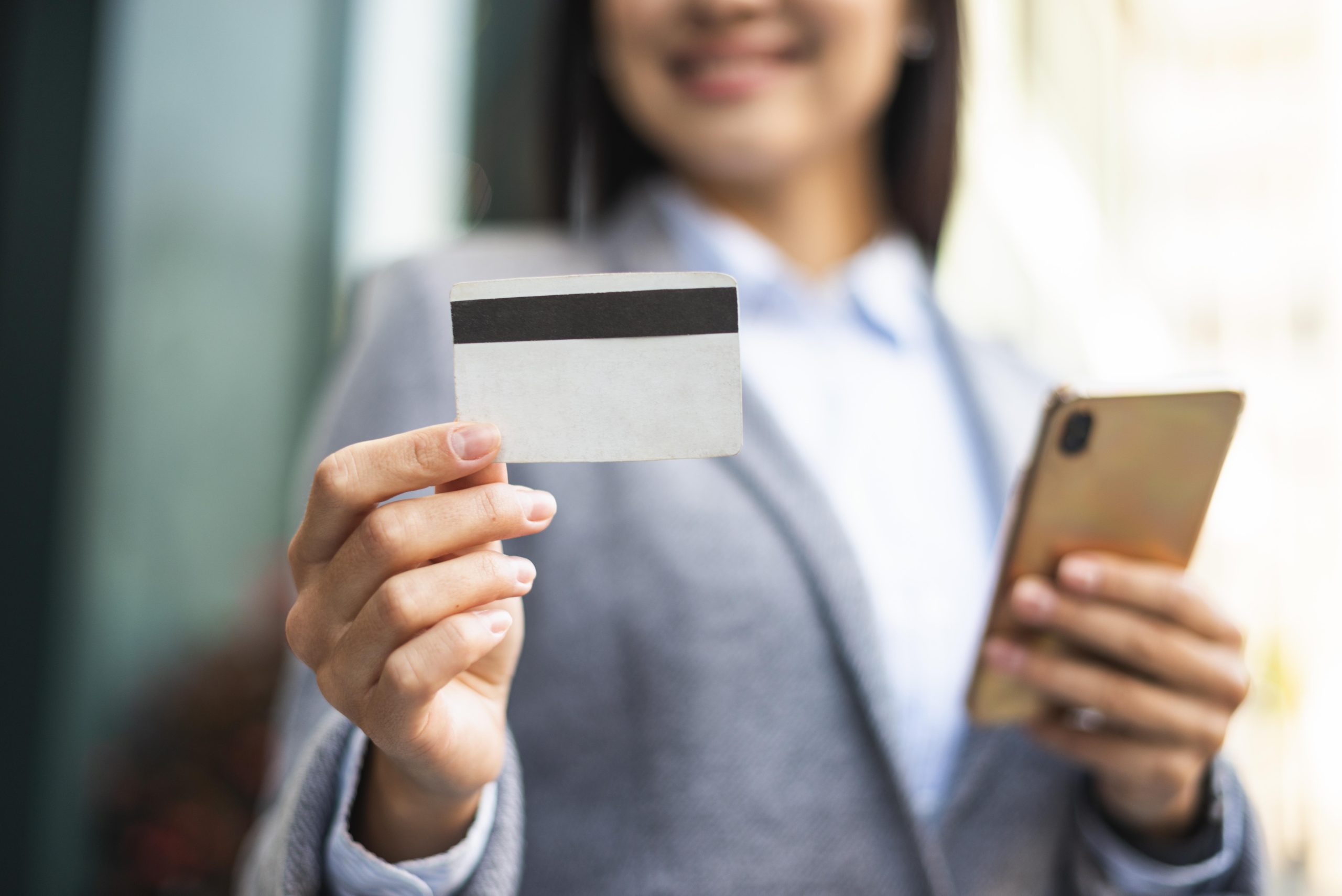A smiling professional woman in a blazer holds a smartphone in one hand and displays a blank credit card in the other. The business setting and confident demeanor highlight the convenience and rewards offered by credit cards with business travel benefits, such as airline miles, hotel perks, and travel insurance.