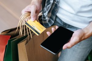 A person holds several shopping bags in one hand along with a yellow credit card, while the other hand holds a smartphone. The scene suggests a recent shopping spree, highlighting the convenience of digital payments. This image represents the use of credit cards for large purchases, emphasizing their practicality and ease in managing high-value transactions.