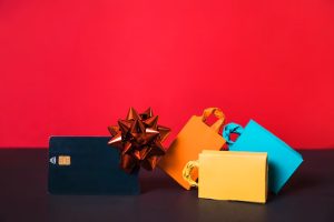 Colorful shopping bags and a wrapped gift card placed on a table against a red background, symbolizing holiday spending and credit cards rewards benefits.