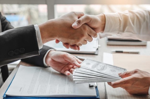 Two business professionals shaking hands over a contract while one hands over dollar bills, representing financial agreements, loans, and the importance of Business Credit in corporate growth.