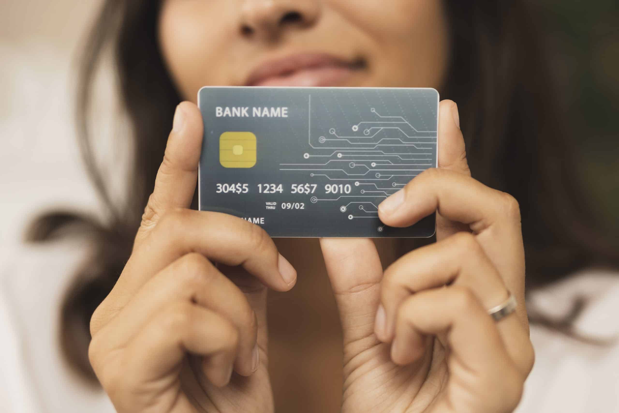 A close-up of a woman holding a credit or debit card between her fingers, displaying it towards the camera. The card has a futuristic design with a dark background, golden chip, and circuit-like patterns. It features placeholder text such as "BANK NAME" and a series of numbers. The woman is slightly out of focus in the background, smiling softly. The image conveys themes of finance, banking, digital transactions, or modern payment methods.