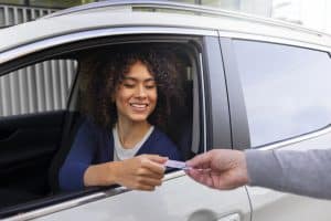 A young woman with curly hair is sitting in the driver's seat of a silver car, smiling as she hands over an identification card to a person outside the vehicle. The person receiving the card is only partially visible, with their hand and a sleeve in focus. The setting appears to be outdoors near a modern building, possibly at a checkpoint or a licensing office.