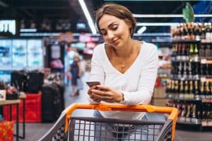 A young woman with short brown hair and light skin is leaning on a shopping cart in a grocery store, smiling while looking at her smartphone. She is wearing a white long-sleeve top, a gold necklace, and a ring on her left hand. The supermarket shelves in the background are stocked with bottles and other products, while a few other shoppers can be seen walking around.