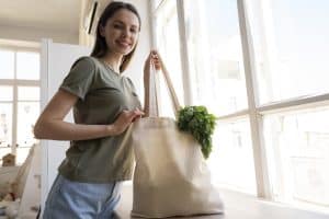 A young woman with long brown hair and light skin is standing near a bright window, smiling at the camera. She is wearing a green t-shirt and light blue jeans. She holds a beige fabric tote bag filled with fresh green herbs, possibly parsley or cilantro, peeking out from the top. The setting appears to be a cozy home with natural light streaming through the large windows, creating a warm and eco-friendly atmosphere.