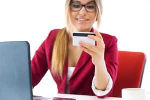 A young woman with long blonde hair, glasses, and light skin is sitting at a desk, holding a credit card while looking at it with a smile. She is wearing a red blazer over a white top. In front of her is an open laptop, and a white coffee cup is placed on the table. The background is bright and minimalistic, suggesting a professional or home office environment. She appears to be making an online purchase or managing her finances.