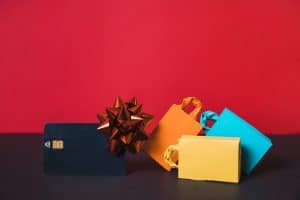 A close-up of a black credit card adorned with an orange gift bow, surrounded by colorful paper shopping bags, against a red background, symbolizing holiday or promotional shopping.