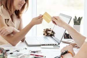 Two people sitting at a desk in an office setting, one handing a gold credit card to the other, with a laptop, documents, keys, and cash visible on the table.