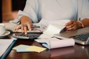 Close-up of a person calculating expenses at a desk with a calculator, laptop, documents, sticky notes, and a cup of coffee.