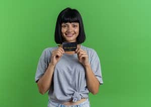 Smiling young woman holding a credit card while standing against a green background, wearing a grey t-shirt and looking at the camera.