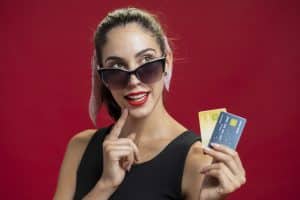 A stylish young woman wearing large black sunglasses and a black sleeveless dress poses confidently against a bold red background. She has a pink scarf tied around her ponytail and bright red lipstick. Holding three colorful high level credit cards in one hand, she gestures thoughtfully with her other hand near her lips, expressing a playful, savvy attitude toward luxury shopping or financial choices.