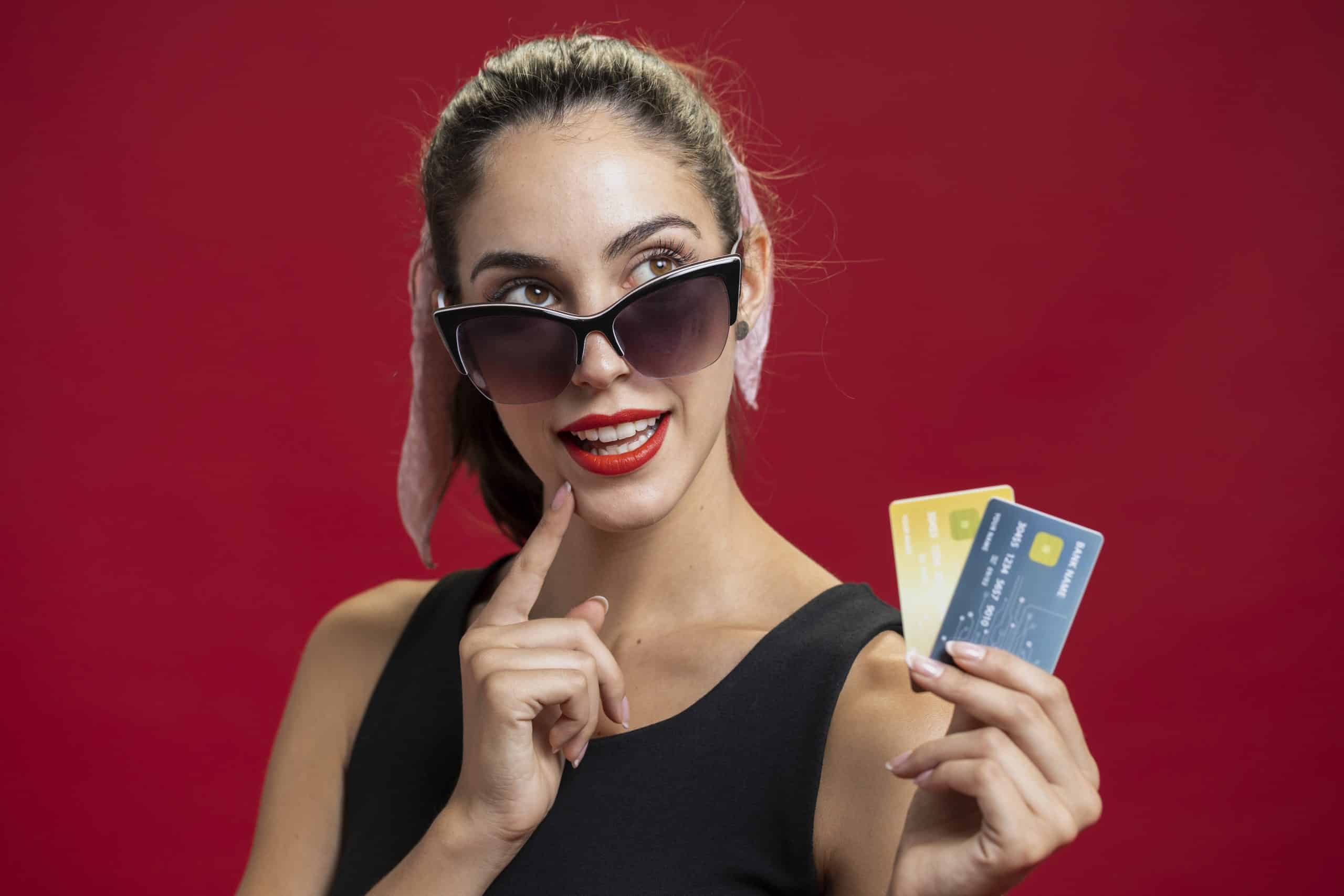 A stylish young woman wearing large black sunglasses and a black sleeveless dress poses confidently against a bold red background. She has a pink scarf tied around her ponytail and bright red lipstick. Holding three colorful high level credit cards in one hand, she gestures thoughtfully with her other hand near her lips, expressing a playful, savvy attitude toward luxury shopping or financial choices.