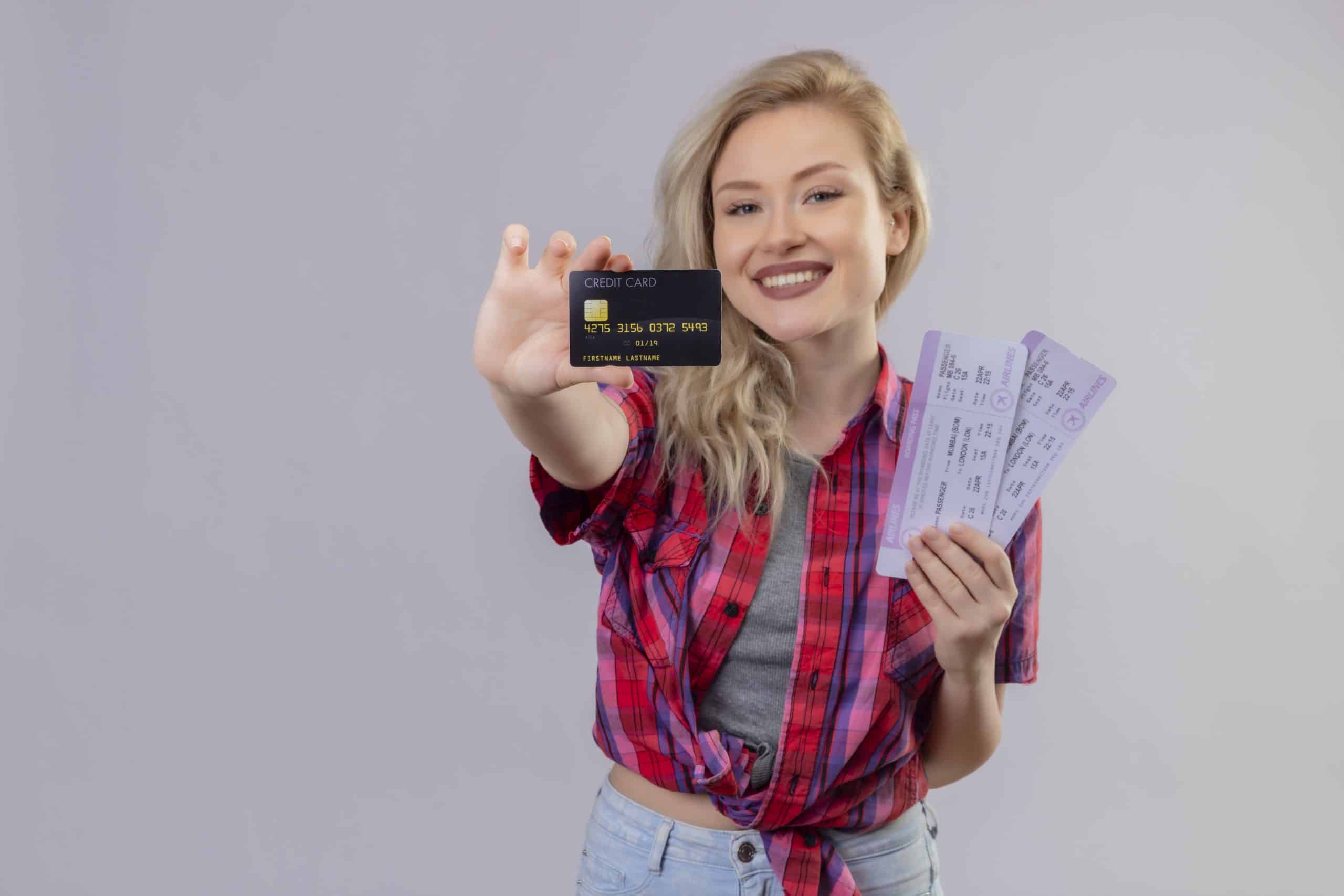 A cheerful young woman with blonde hair smiles while holding a black credit card in one hand and two airline boarding passes in the other. She is dressed casually in a red plaid shirt and light denim jeans, standing against a plain light background. This image visually represents the travel perks and rewards made possible by cashback credit cards, emphasizing convenience and financial benefit.