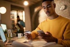 A focused young man in a mustard yellow sweatshirt sits at a desk in a modern workspace, holding a credit card in one hand and a smartphone in the other. Surrounded by laptops, documents, and tech accessories, he reviews information attentively, suggesting online financial management. This image illustrates the practical use of credit card benefits for technology professionals balancing efficiency and digital tools.