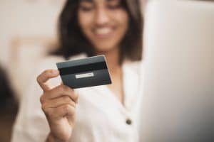 A smiling woman holds a credit card while sitting in front of a laptop, symbolizing access to digital banking tools. She wears a white blouse and appears engaged in an online transaction, highlighting the ease and accessibility of modern financial services. The image represents the growing role of credit cards in the financial inclusion of underserved or emerging consumer groups.