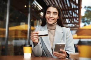 A confident young woman sits at an outdoor café, smiling while holding a smartphone in one hand and a credit card in the other. She wears a stylish light gray coat over a beige turtleneck sweater, with a takeaway coffee cup placed on the wooden table in front of her. The image reflects modern financial independence and the growing use of credit cards for entrepreneurs managing business on the go.