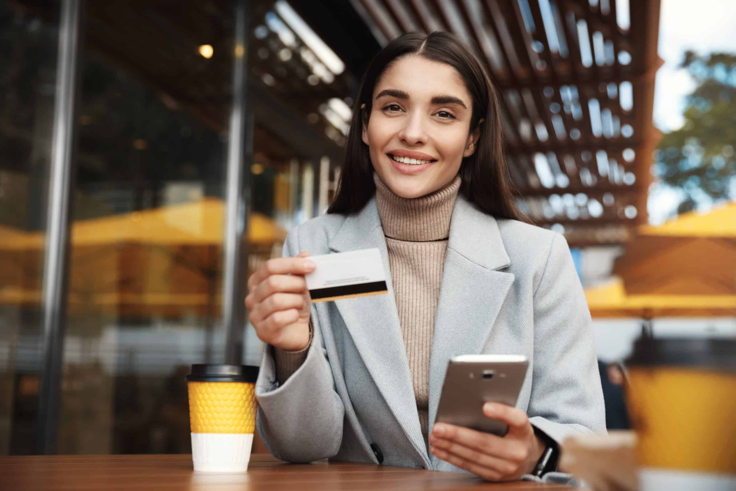 A confident young woman sits at an outdoor café, smiling while holding a smartphone in one hand and a credit card in the other. She wears a stylish light gray coat over a beige turtleneck sweater, with a takeaway coffee cup placed on the wooden table in front of her. The image reflects modern financial independence and the growing use of credit cards for entrepreneurs managing business on the go.