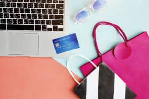 A flat lay of a laptop keyboard, a blue credit card labeled "PREMIUM Credit," colorful shopping bags, and sunglasses on a pastel background. The vibrant setup represents the ease and convenience of using credit cards for international shopping, combining online access with global style.