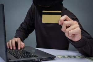 A masked individual in dark clothing uses a laptop while holding up a gold credit card, with dollar bills visible on the table. The image highlights cybersecurity threats and fraud risks associated with credit cards in the digital age.