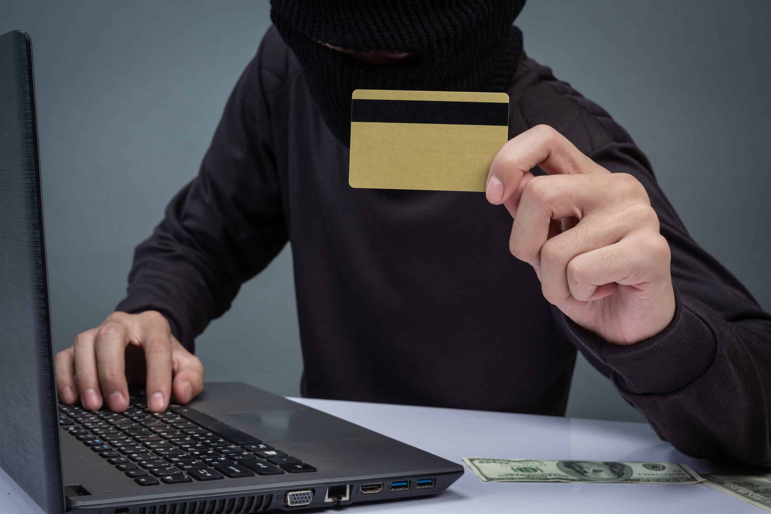 A masked individual in dark clothing uses a laptop while holding up a gold credit card, with dollar bills visible on the table. The image highlights cybersecurity threats and fraud risks associated with credit cards in the digital age.