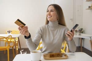 A cheerful woman sits at a café table with a slice of cake and a cup of coffee, holding a gold credit card in one hand and a smartphone in the other. Her relaxed and happy demeanor reflects the lifestyle advantages offered by credit cards with wellness benefits, such as rewards for dining, relaxation, and self-care experiences.