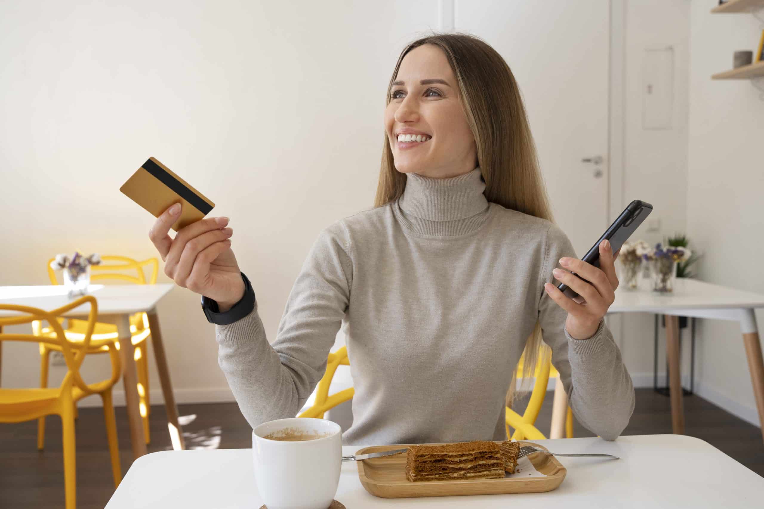 A cheerful woman sits at a café table with a slice of cake and a cup of coffee, holding a gold credit card in one hand and a smartphone in the other. Her relaxed and happy demeanor reflects the lifestyle advantages offered by credit cards with wellness benefits, such as rewards for dining, relaxation, and self-care experiences.