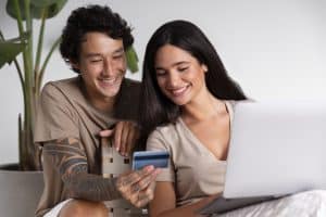 A smiling millennial couple sits together indoors, engaged in online shopping. The woman holds a laptop on her lap while the man shows her a credit card, both appearing relaxed and happy. He has tribal tattoos on his arm, and they are casually dressed in neutral tones. This scene captures the digital-savvy lifestyle of millennials using technology and credit cards for shared financial decisions.