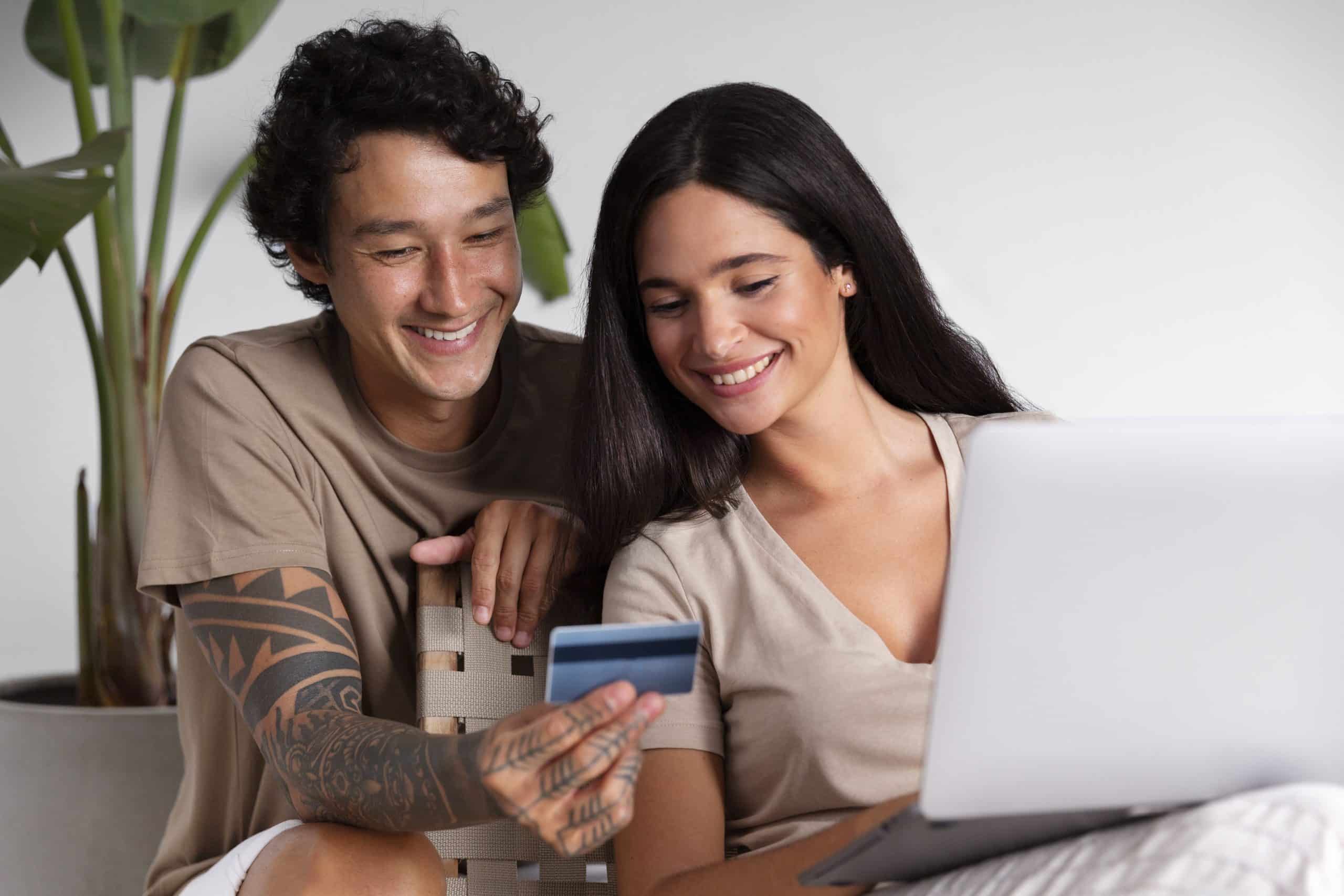 A smiling millennial couple sits together indoors, engaged in online shopping. The woman holds a laptop on her lap while the man shows her a credit card, both appearing relaxed and happy. He has tribal tattoos on his arm, and they are casually dressed in neutral tones. This scene captures the digital-savvy lifestyle of millennials using technology and credit cards for shared financial decisions.