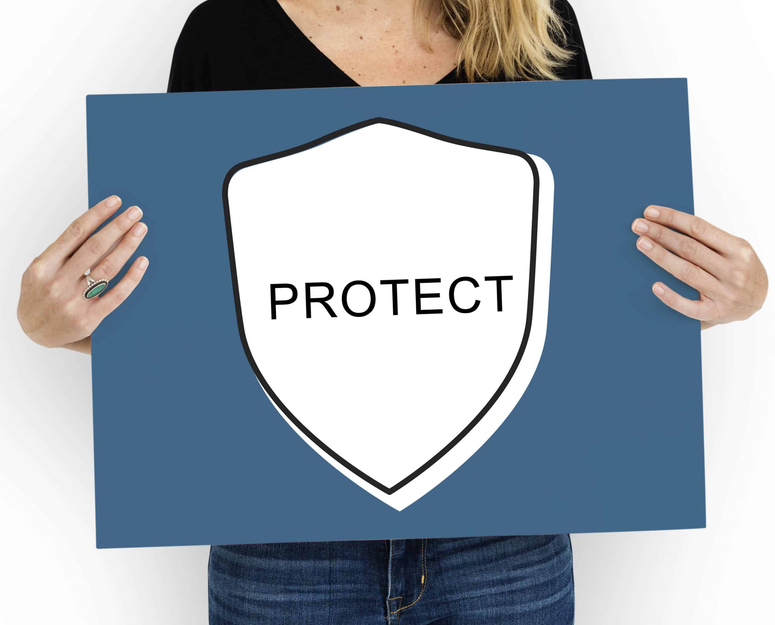 A woman holds a blue sign with a white shield icon in the center, outlined in black. Inside the shield, the word “PROTECT” is written in bold black letters, symbolizing safety and defense. This image visually represents the concept of new consumer protection rules aimed at safeguarding buyer rights and promoting responsible practices.