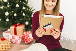 A smiling woman with red-painted nails uses a tablet and holds a credit card while sitting in front of a beautifully decorated Christmas tree surrounded by wrapped gifts. She wears a cozy burgundy sweater and appears to be enjoying online seasonal shopping. The festive background and digital device emphasize the ease and popularity of using a credit card for holiday purchases.
