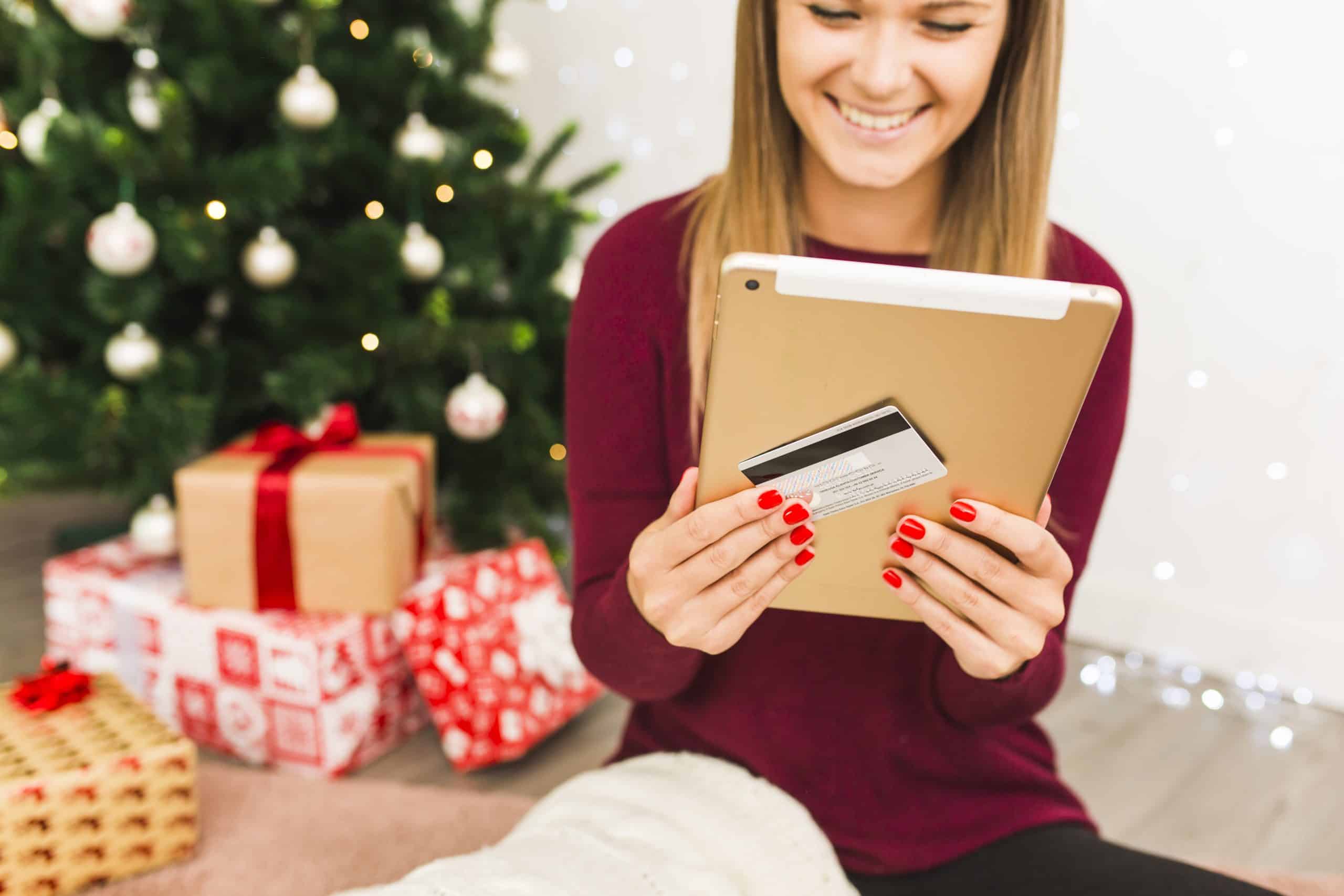 A smiling woman with red-painted nails uses a tablet and holds a credit card while sitting in front of a beautifully decorated Christmas tree surrounded by wrapped gifts. She wears a cozy burgundy sweater and appears to be enjoying online seasonal shopping. The festive background and digital device emphasize the ease and popularity of using a credit card for holiday purchases.
