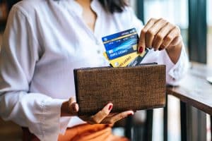 A woman wearing a white blouse and with neatly manicured red nails pulls multiple credit cards from a brown textured wallet while seated at a wooden table in a well-lit indoor setting. The cards include gold and blue tones, symbolizing status and financial power. This image visually conveys the concept of a wealth credit card used for premium financial management and exclusive benefits.