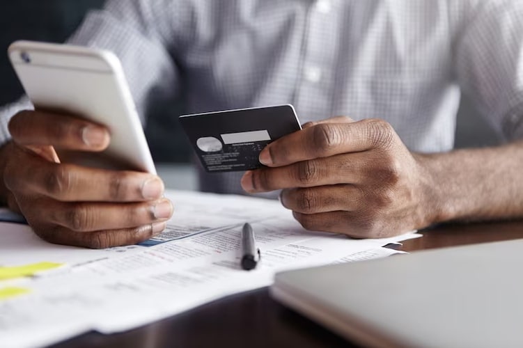 A person holding a smartphone and a black credit card while reviewing financial documents at a desk, illustrating the role of credit cards in managing personal expenses.