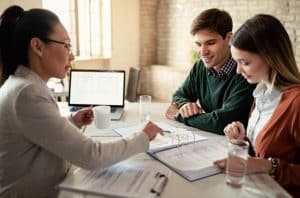 A couple sitting at a desk with a financial advisor, reviewing documents and blueprints together. A laptop and glasses of water are on the table, suggesting a discussion about personal loans and financial planning.