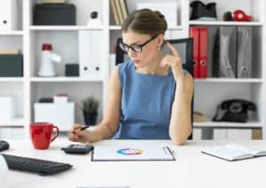 A focused businesswoman sitting at her desk analyzing a credit score chart with a calculator in hand, surrounded by office supplies and documents. The image reflects financial planning or loan evaluation possibly related to SBA (Small Business Administration) support for small businesses.