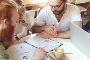 A group of young professionals sitting at a table in a casual workspace, reviewing charts and financial documents together with highlighters and a laptop. The image represents a collaborative planning session focused on financing for startups.