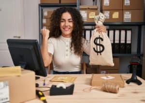 A smiling woman working in a small business holds a money bag and cash at her desk, representing how minority businesses are accessing credit to support growth and operations.