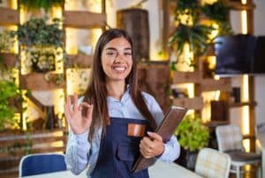 A smiling businesswoman in an apron holding a folder and making an OK gesture, symbolising confidence and success in securing franchise expansion for loans.