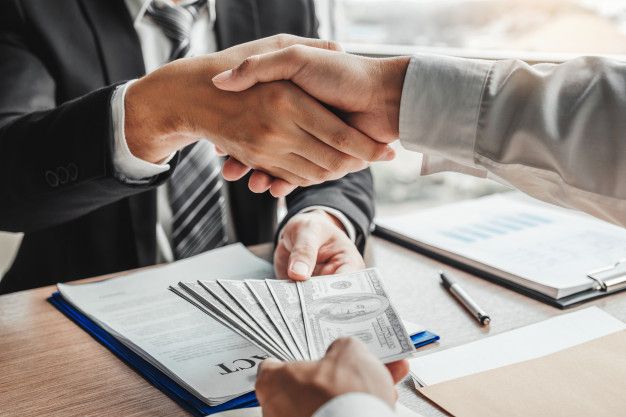 Businessmen shaking hands over a desk while exchanging cash, symbolising financial agreements and access to loans for immigrants.